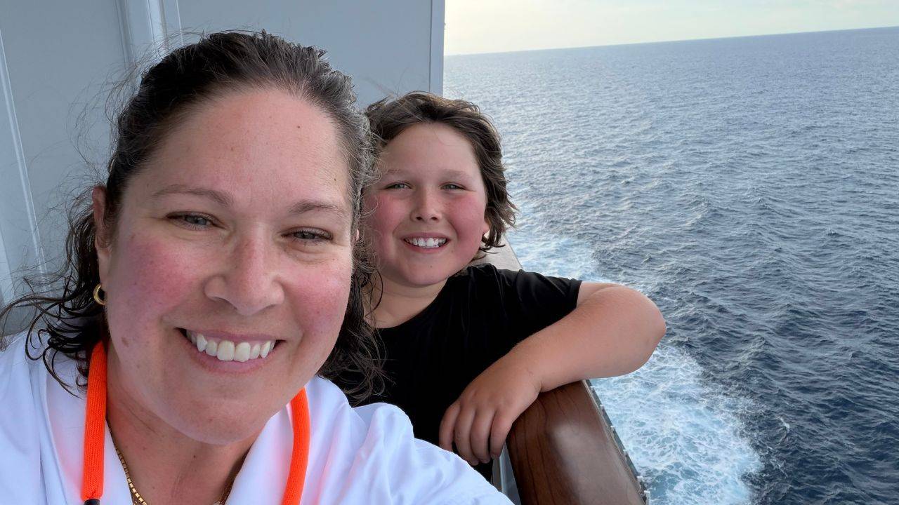 A woman and young boy smile for a selfie on the side of a cruise ship, with the open ocean and the ship’s wake stretching behind them.