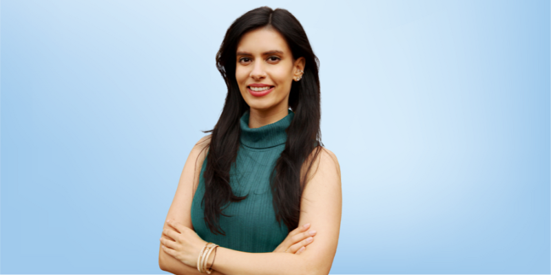 Anya Charles, Energy Magazine Editor-in-Chief for the Healing Touch Program, standing with arms crossed against a light blue background.