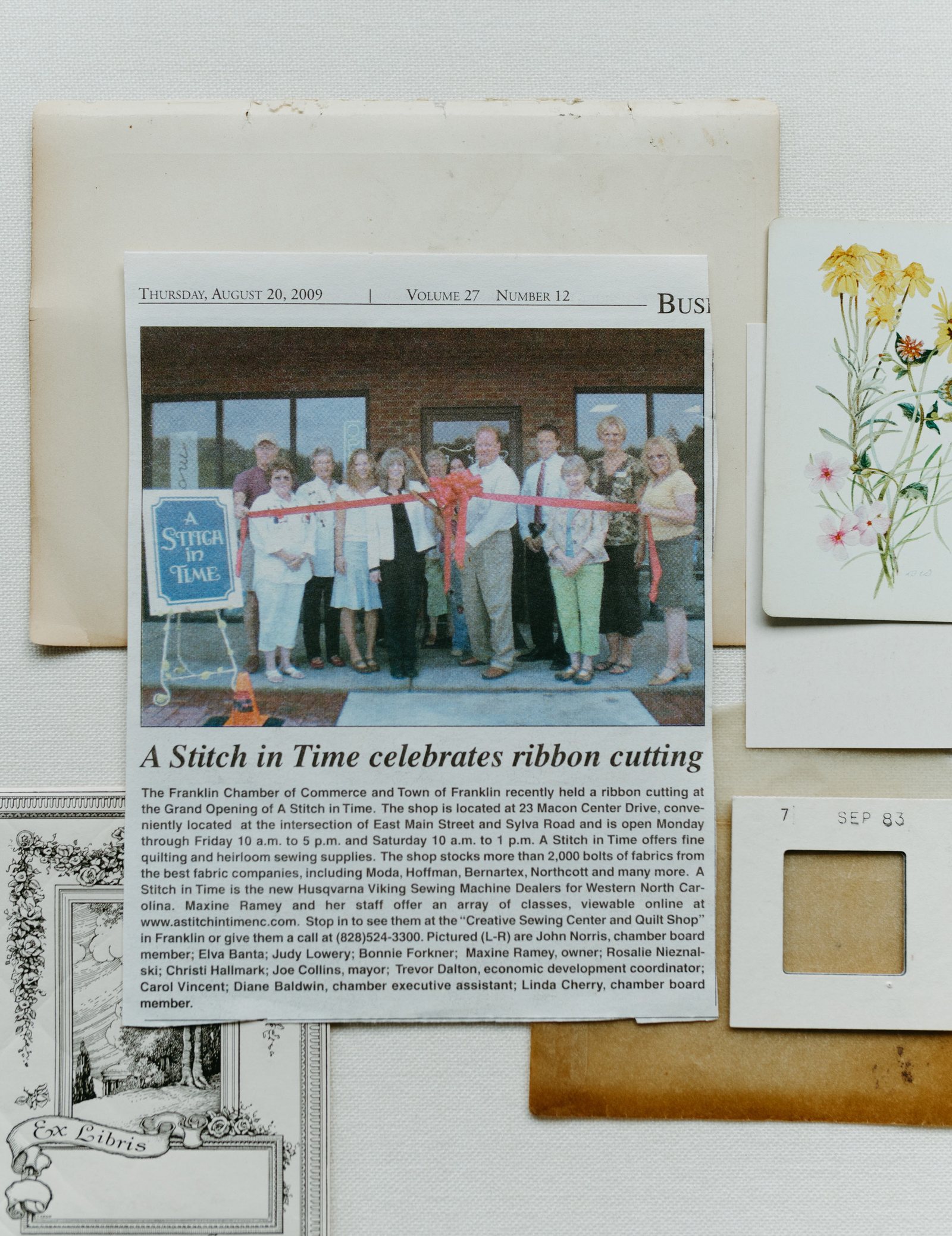 A 2009 newspaper clipping of Bonnie Christine at A Stitch in Time’s ribbon cutting, laid among vintage papers and a botanical card