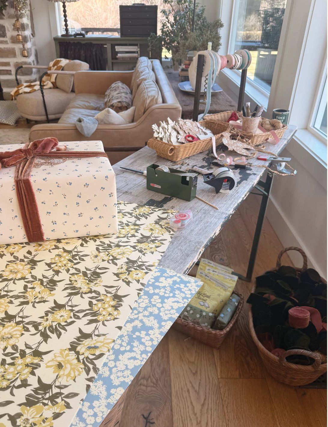 The wrapping station: a wooden table by the window stacked with patterned paper and wrapped gifts, the living room beyond