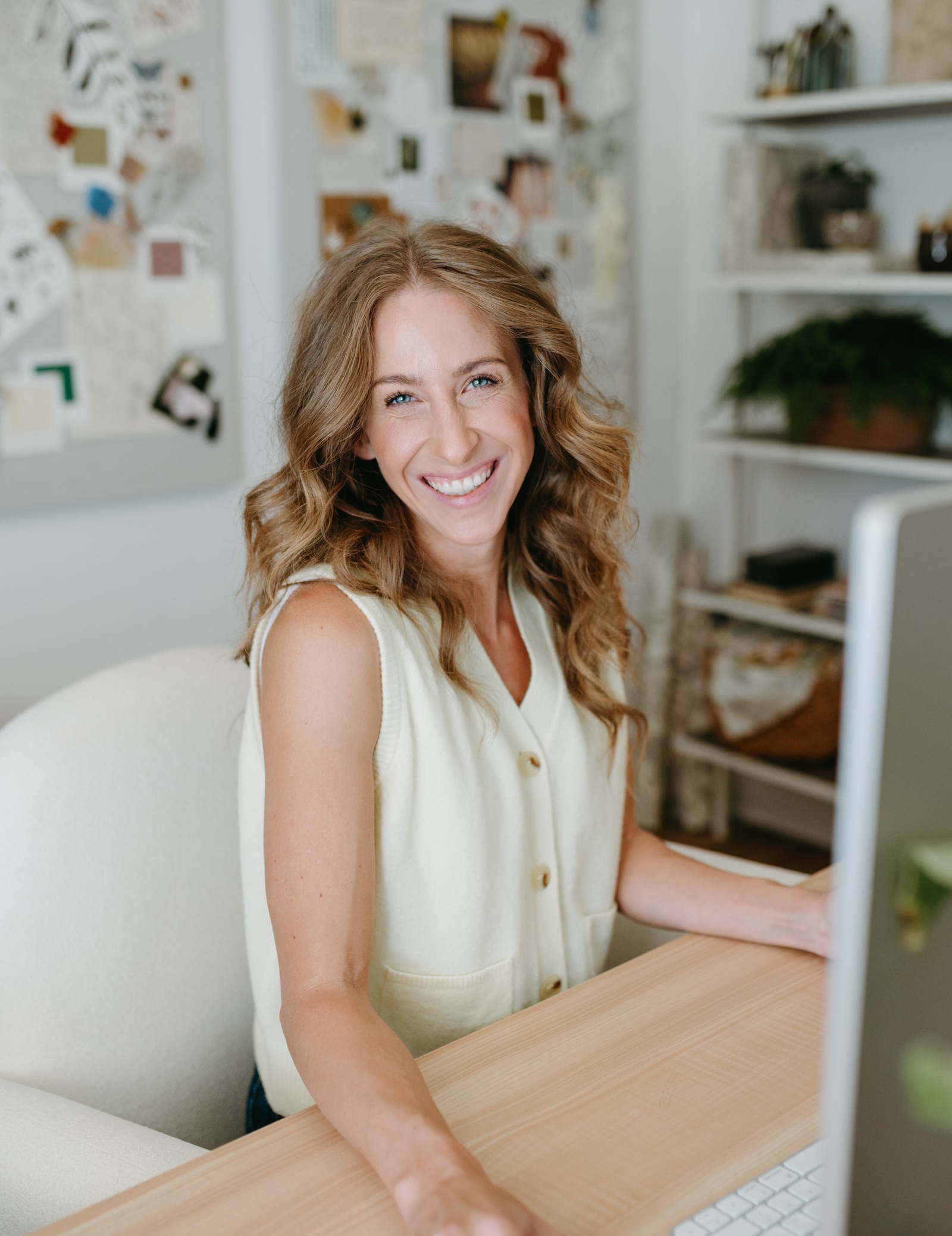 Bonnie Christine typing at a wooden desk in her studio, a mood board on the wall