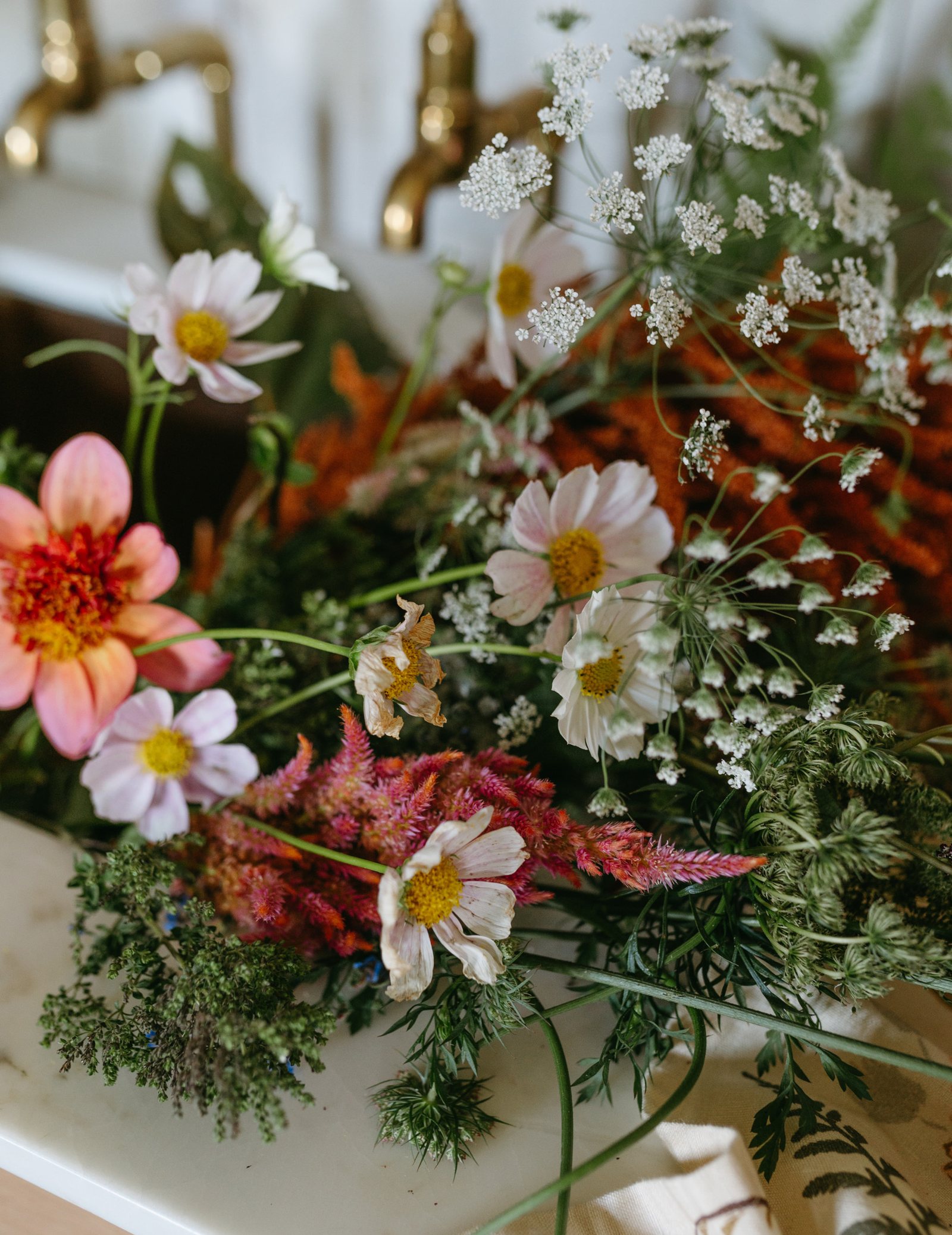 Pink dahlias, cosmos, and wildflowers gathered in a farmhouse sink beneath brass taps