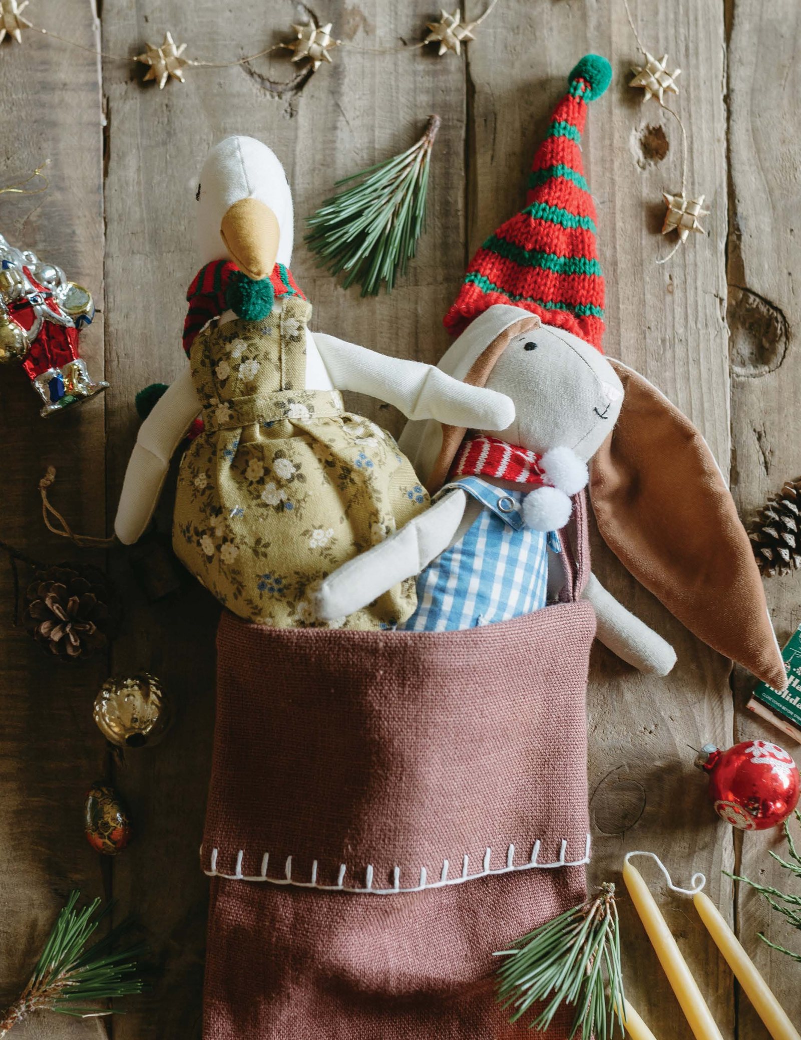 Two Flowerie softie dolls in holiday hats tucked into a linen stocking, with pine sprigs and ornaments