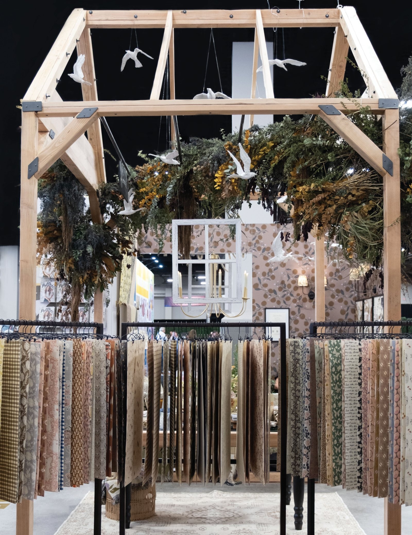 The wooden greenhouse booth at the Licensing Expo, with dried florals over the beams, paper birds overhead, and fabric swatches hanging in rows