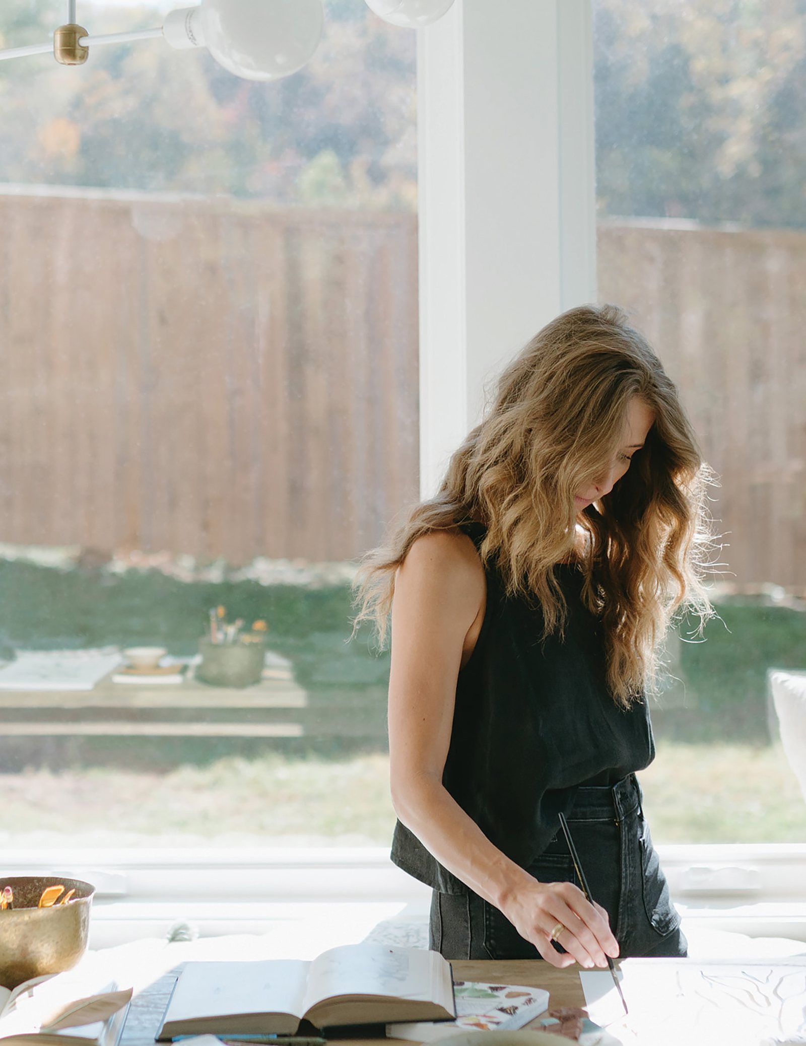 Bonnie Christine at her studio table by a tall window, opening a sketchbook