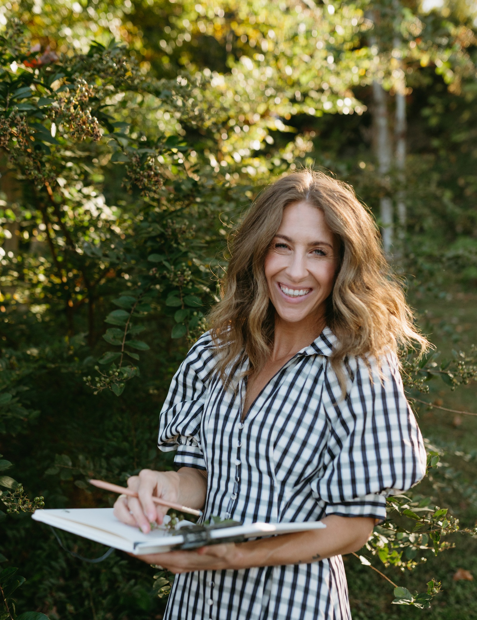 Bonnie Christine laughing in her garden in a gingham dress, holding a sketchbook and pencil