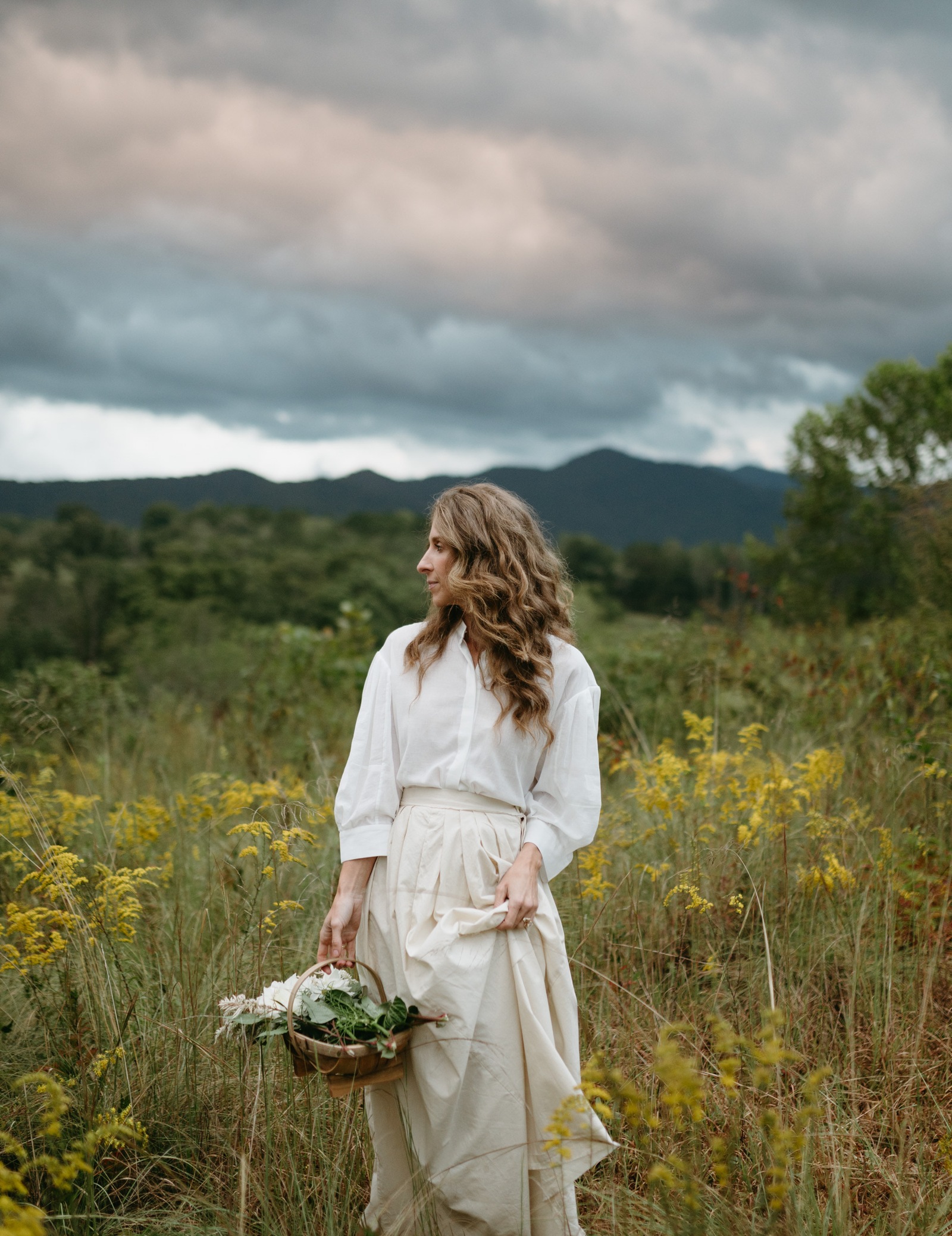 Bonnie Christine walking through a meadow of goldenrod with a basket of flowers, mountains and storm clouds behind her