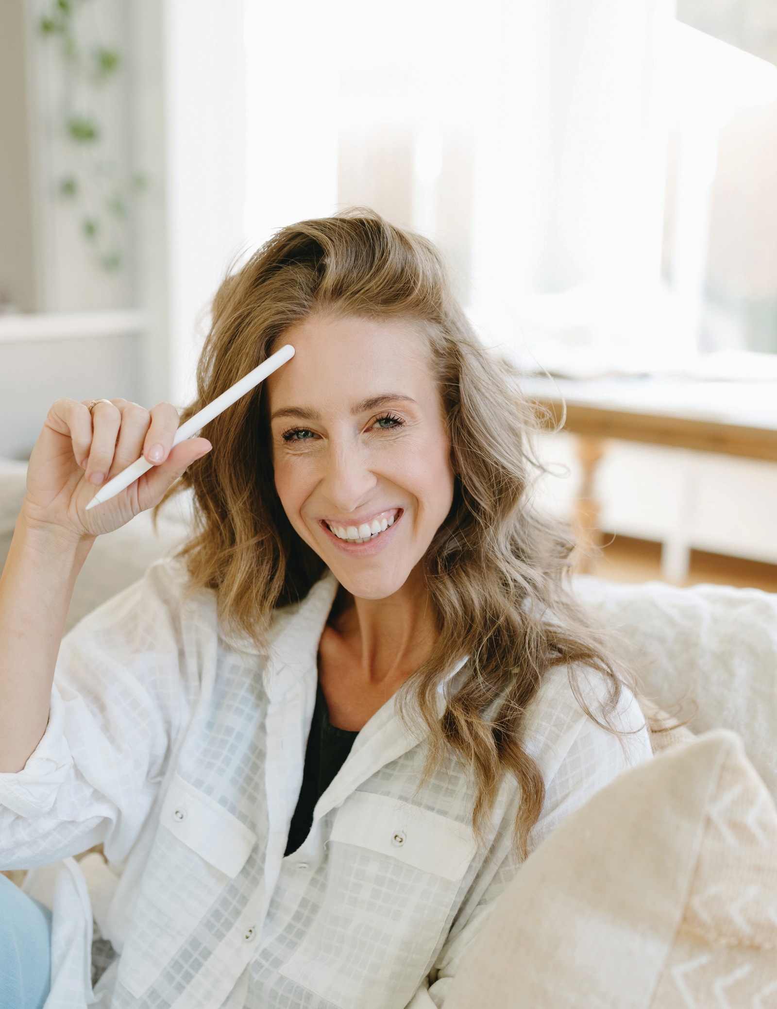 Bonnie Christine smiling on a sofa in her bright studio, holding a white digital stylus up near her forehead