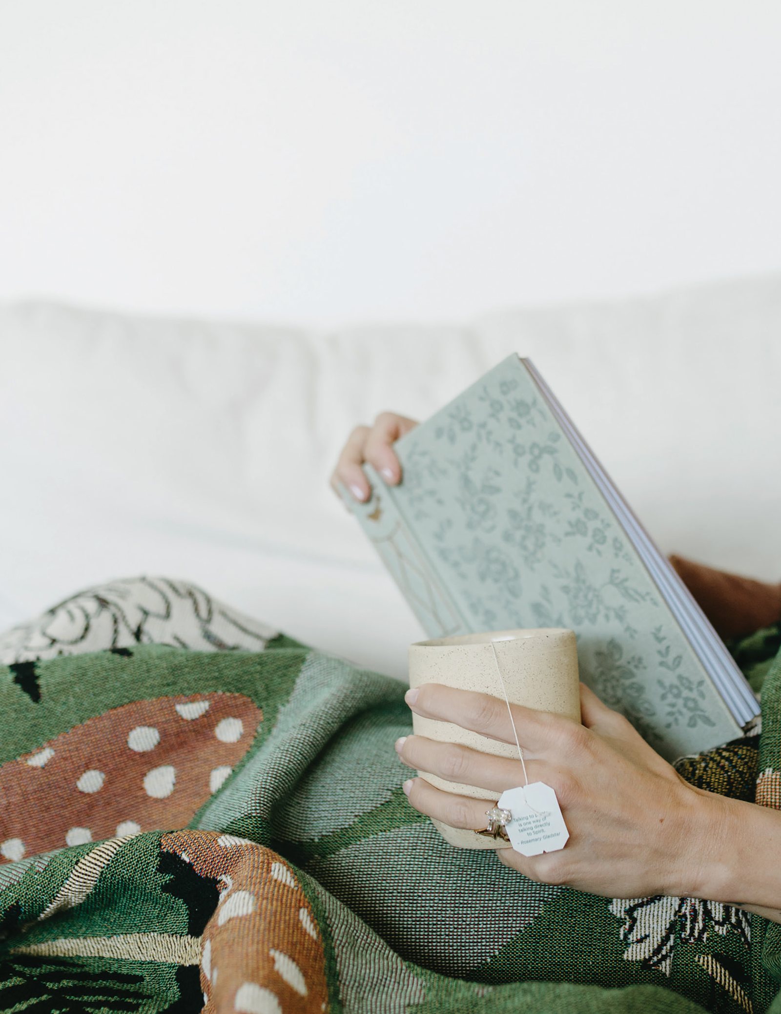 A hand holding a mug of coffee beside a sage green book, curled up under a blanket
