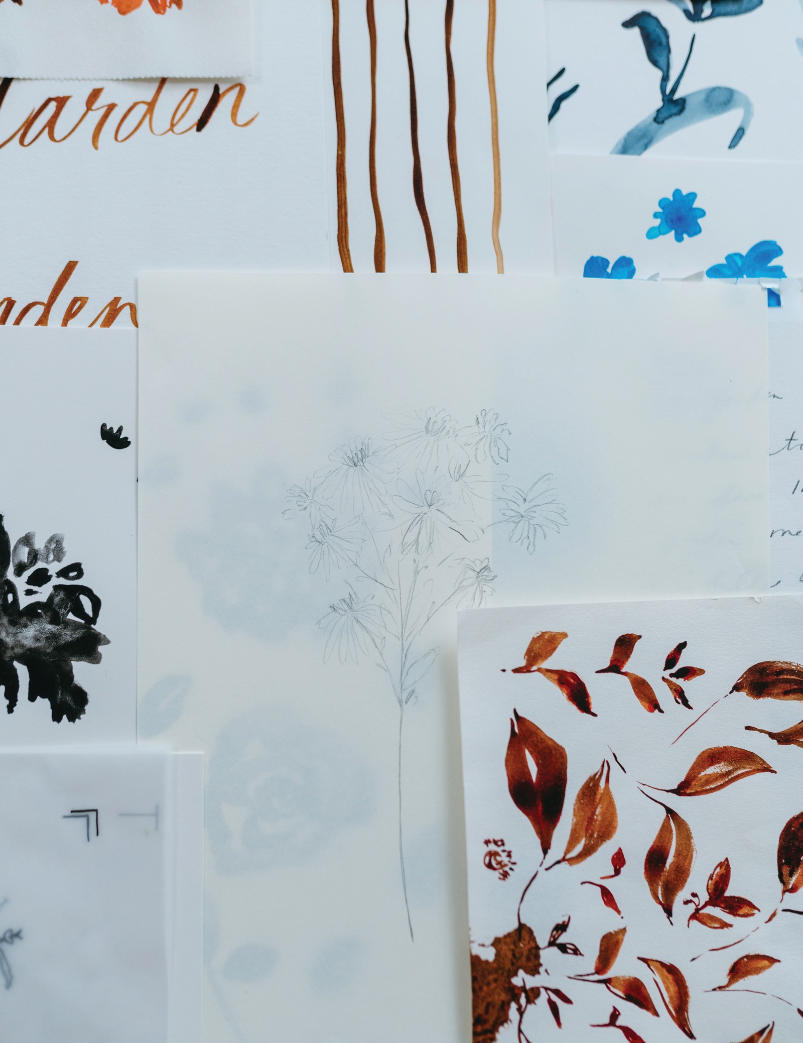 Watercolor and pencil studies layered on a wall: “Garden” lettering, rust leaves, blue florals, and a pencil flower sketch