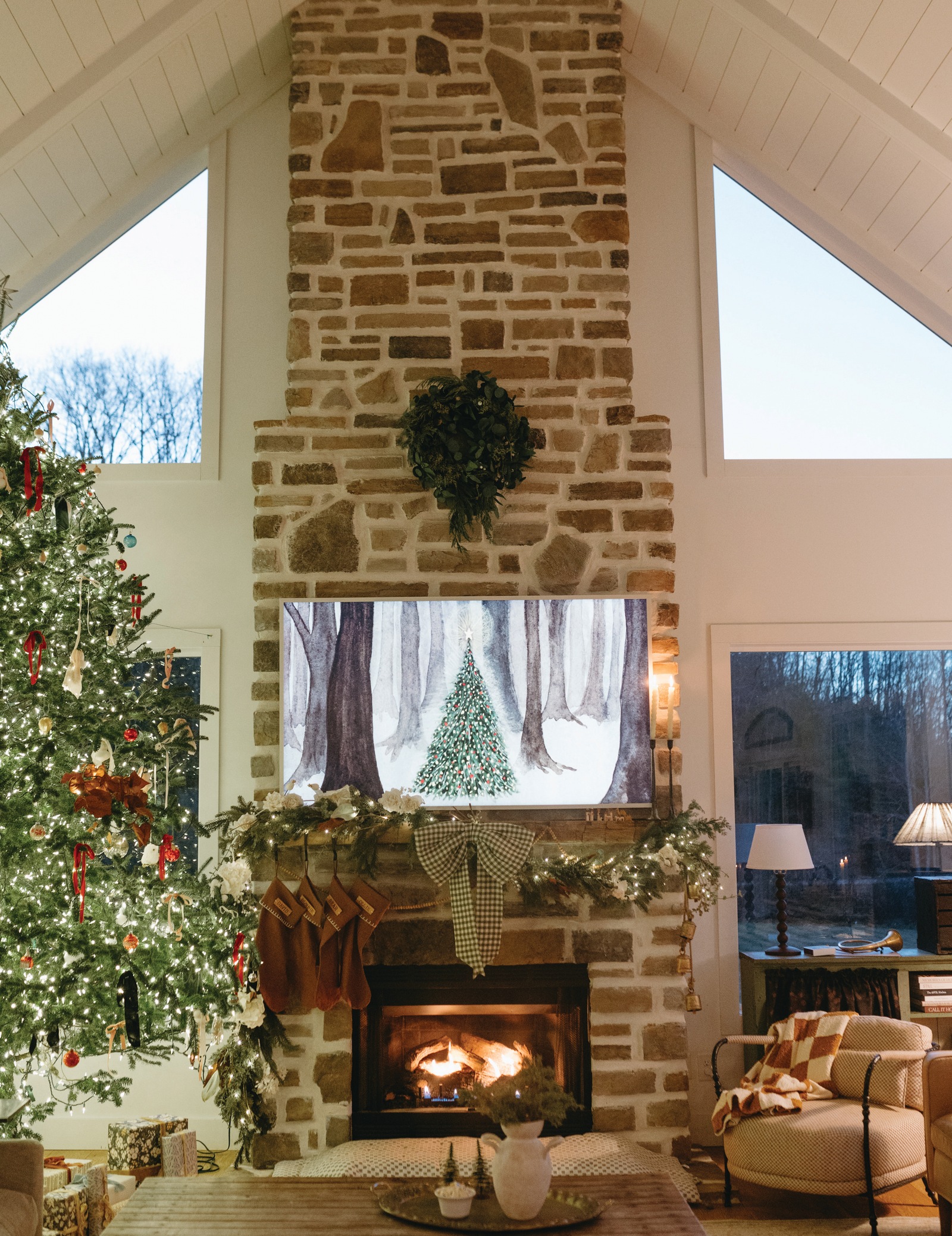 A cozy living room at Christmas with a lit tree beside a tall stone fireplace, stockings and garland on the mantel, and a fire glowing