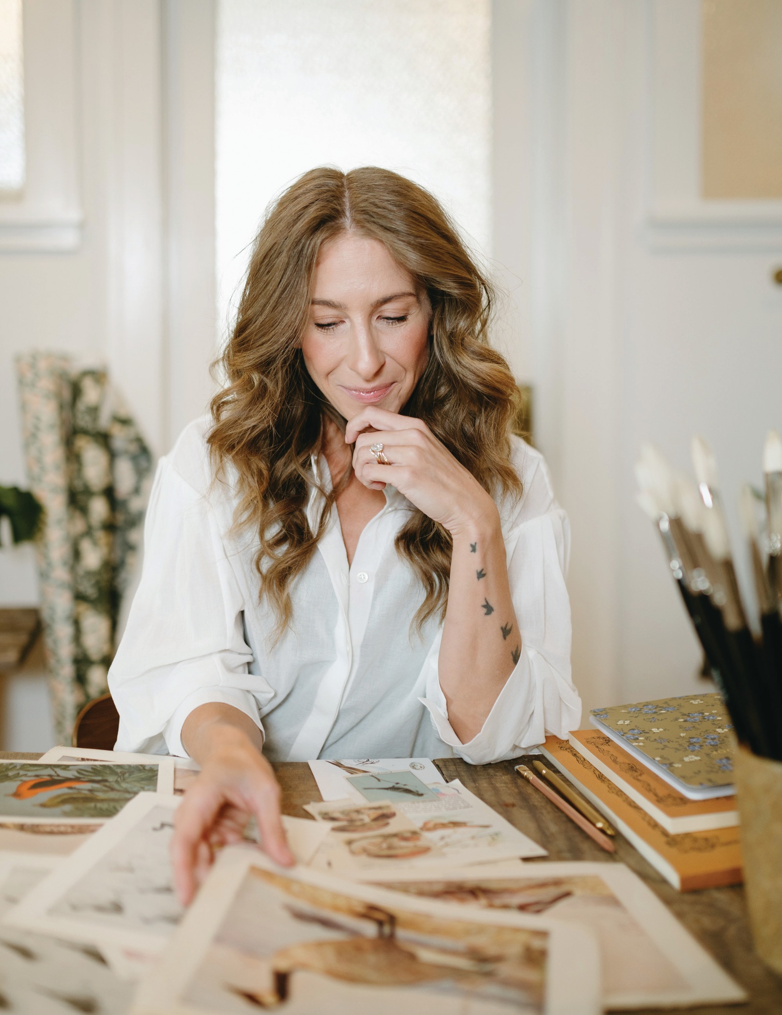 Bonnie Christine smiling at her studio table as she looks through vintage bird prints, with notebooks and paintbrushes beside her