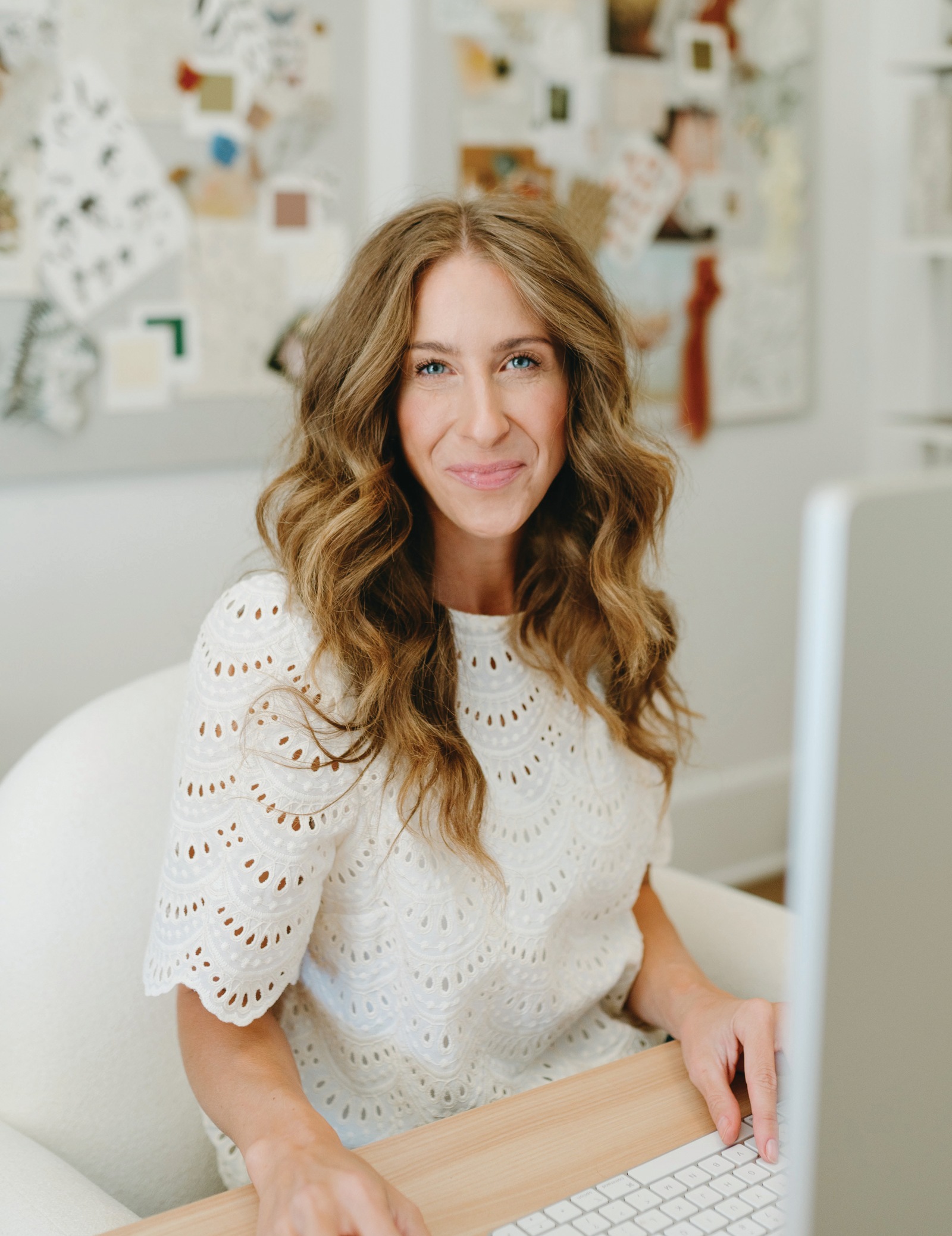 Bonnie Christine smiling at her desk in a white eyelet top, with inspiration boards on the studio wall behind her