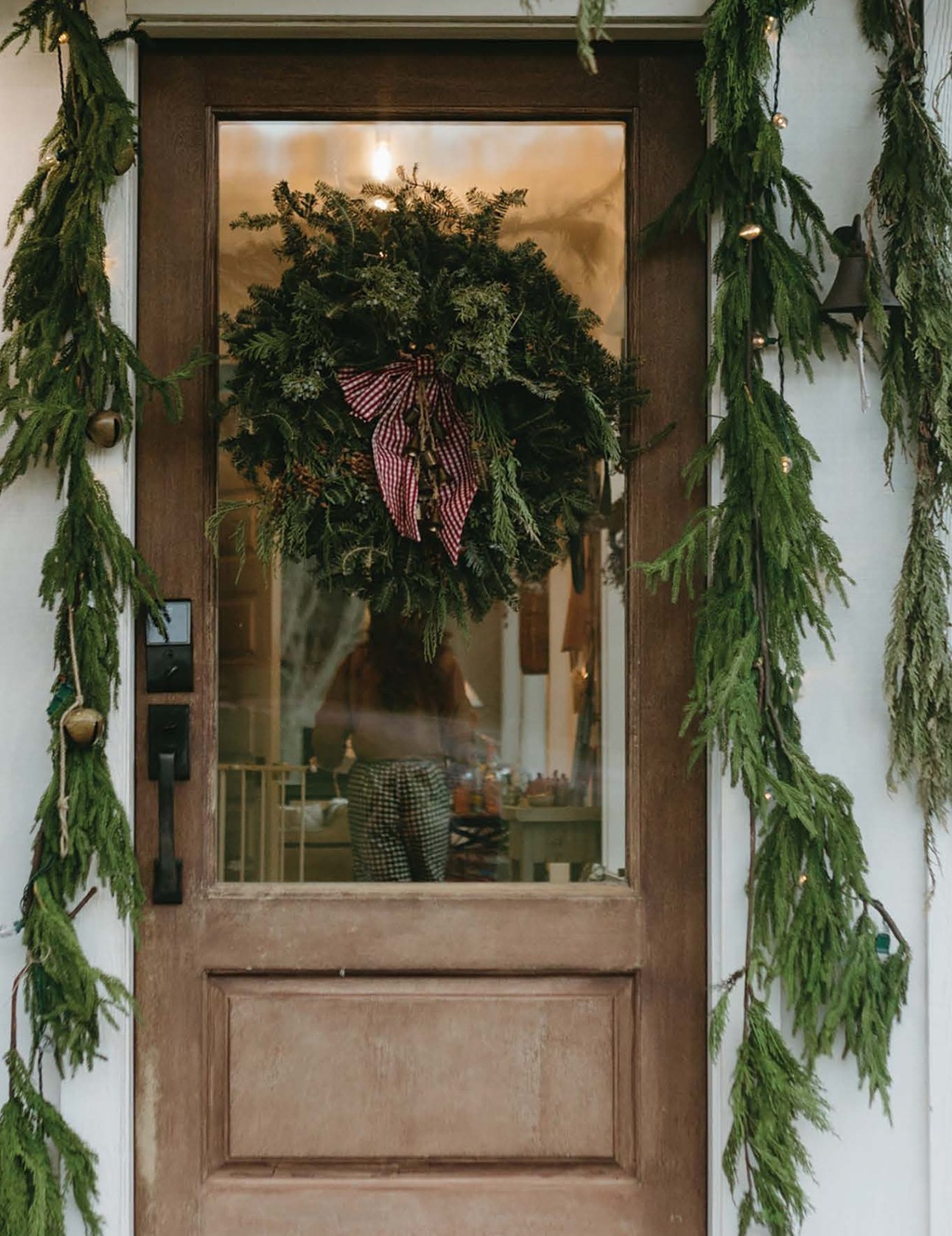 A wooden front door hung with a wreath and cedar garland, lanterns lit on either side