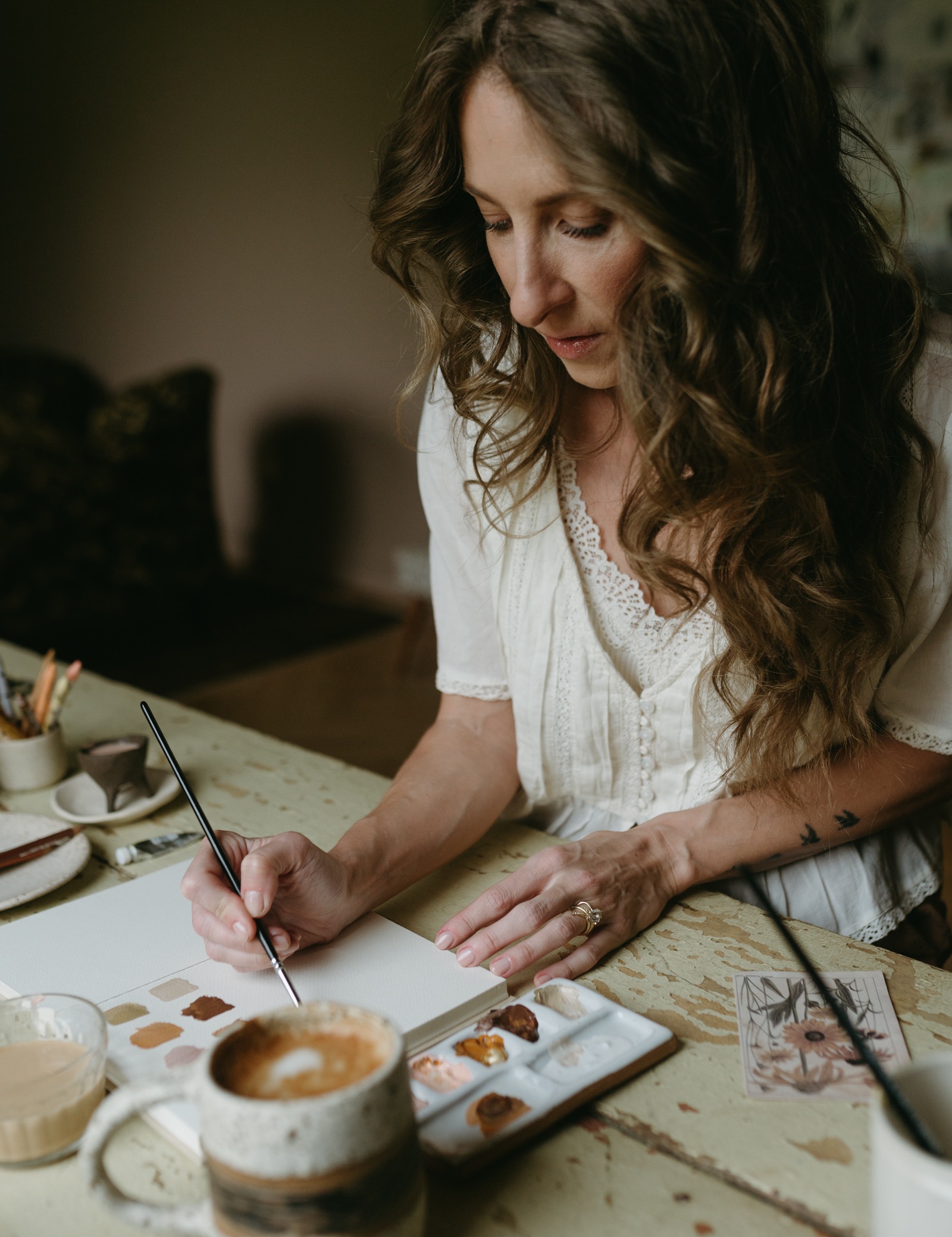 Bonnie Christine painting at a worn wooden table in her analog studio, a latte beside her