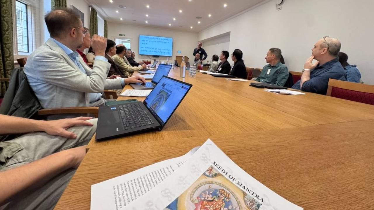 Scholars seated around a long conference table at Harris Manchester College, Oxford, listening to a presentation with Sanskrit text on the screen, a Seeds of Mantra handout in the foreground