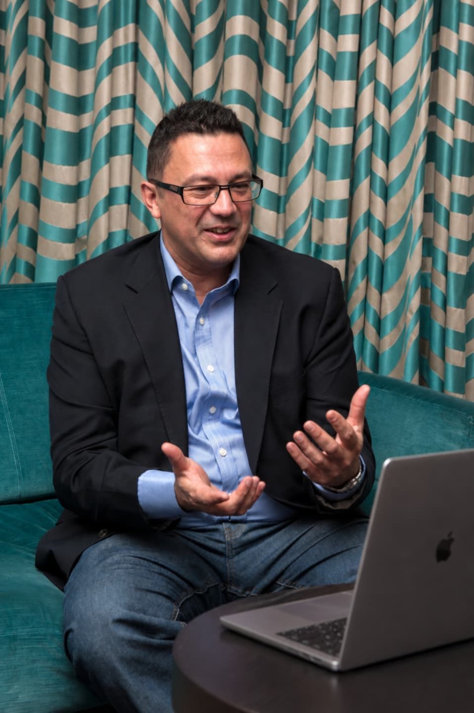Roy Ripper sitting on a teal sofa during an online interview or coaching session, speaking with hand gestures beside an open laptop.