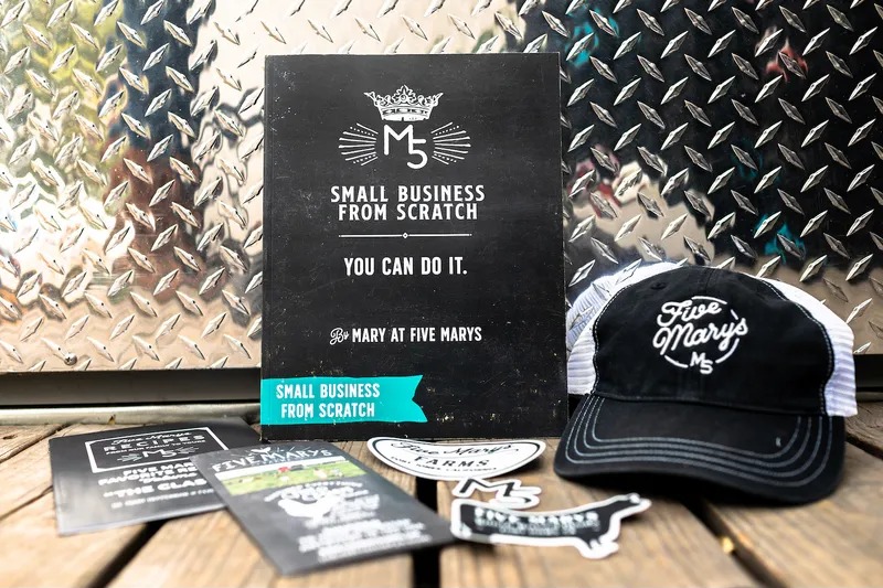 The original Small Business From Scratch workbook by Mary at Five Marys, standing next to a Five Marys cap
