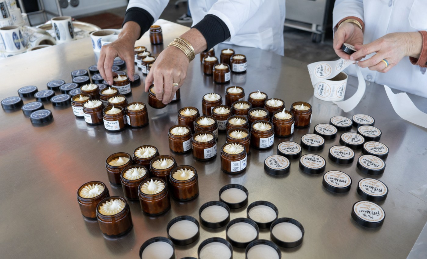 Hands labeling jars of whipped Five Marys tallow on a steel table