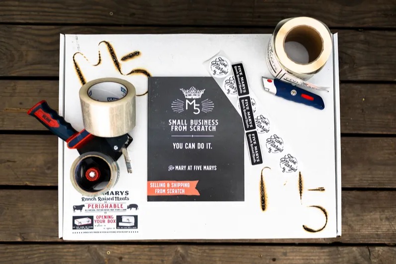 The original Selling & Shipping From Scratch workbook on a Five Marys shipping box with packing tape and labels