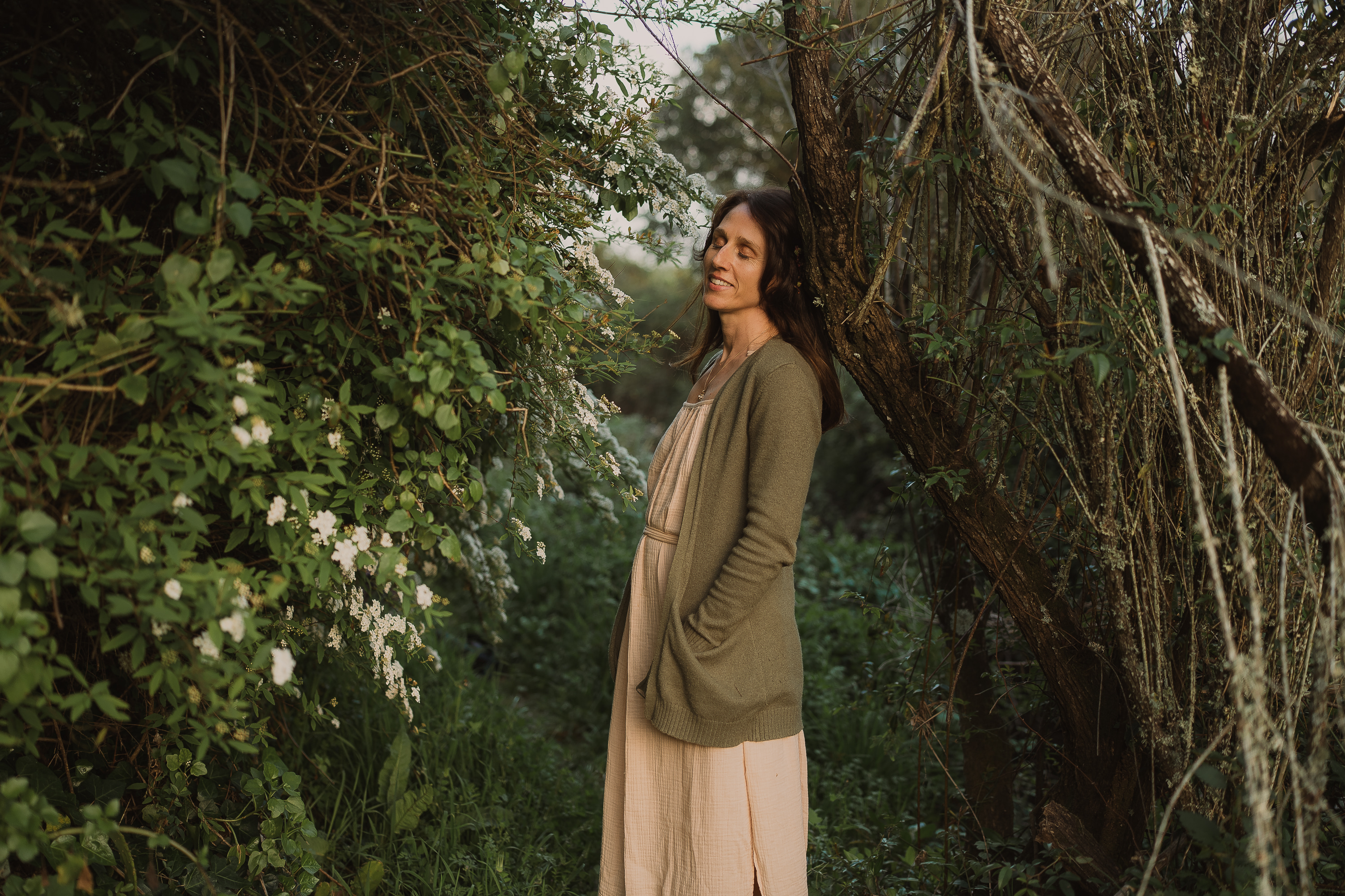 Image: Amelia standing in dense forest undergrowth, eyes closed, leaning against wild branching trees and white flowers, green cardigan, golden hour 