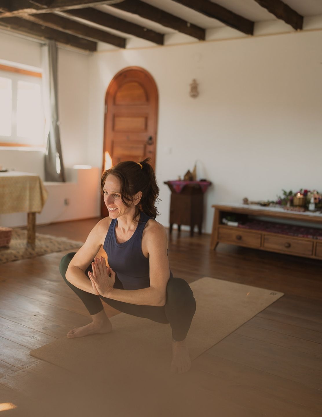 Image: Amelia in yoga squat pose on mat, Portugal studio, wooden beams, altar visible in background, smiling, joyful