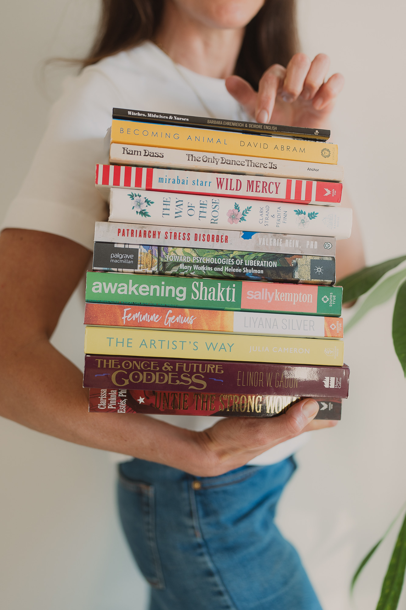  Image: Amelia holding a large stack of books with visible spines — David Abram, Mirabai Starr, Patriarchy Stress Disorder, Awakening Shakti, The Artist's Way, Clarissa Pinkola Estés among others 