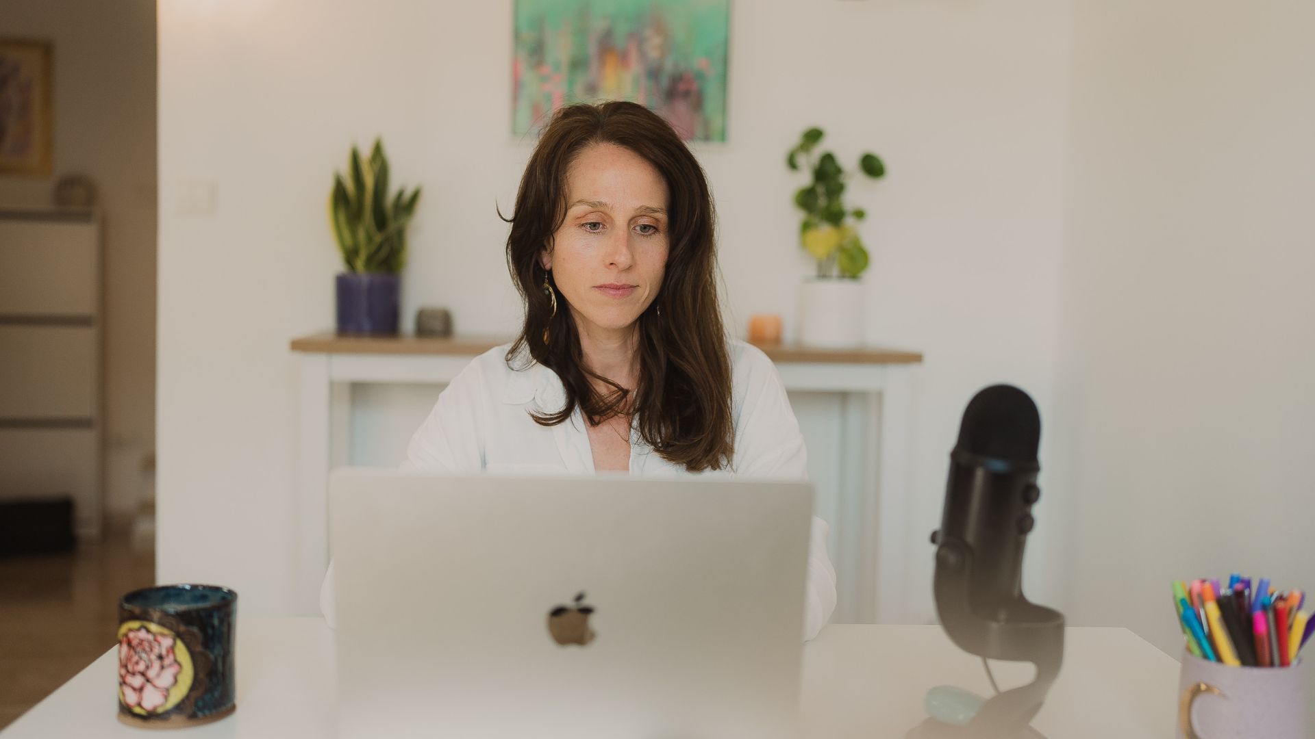 Spiritual business coach Amelia Travis sits at her computer, in office with natural light and green plants, on a client call.