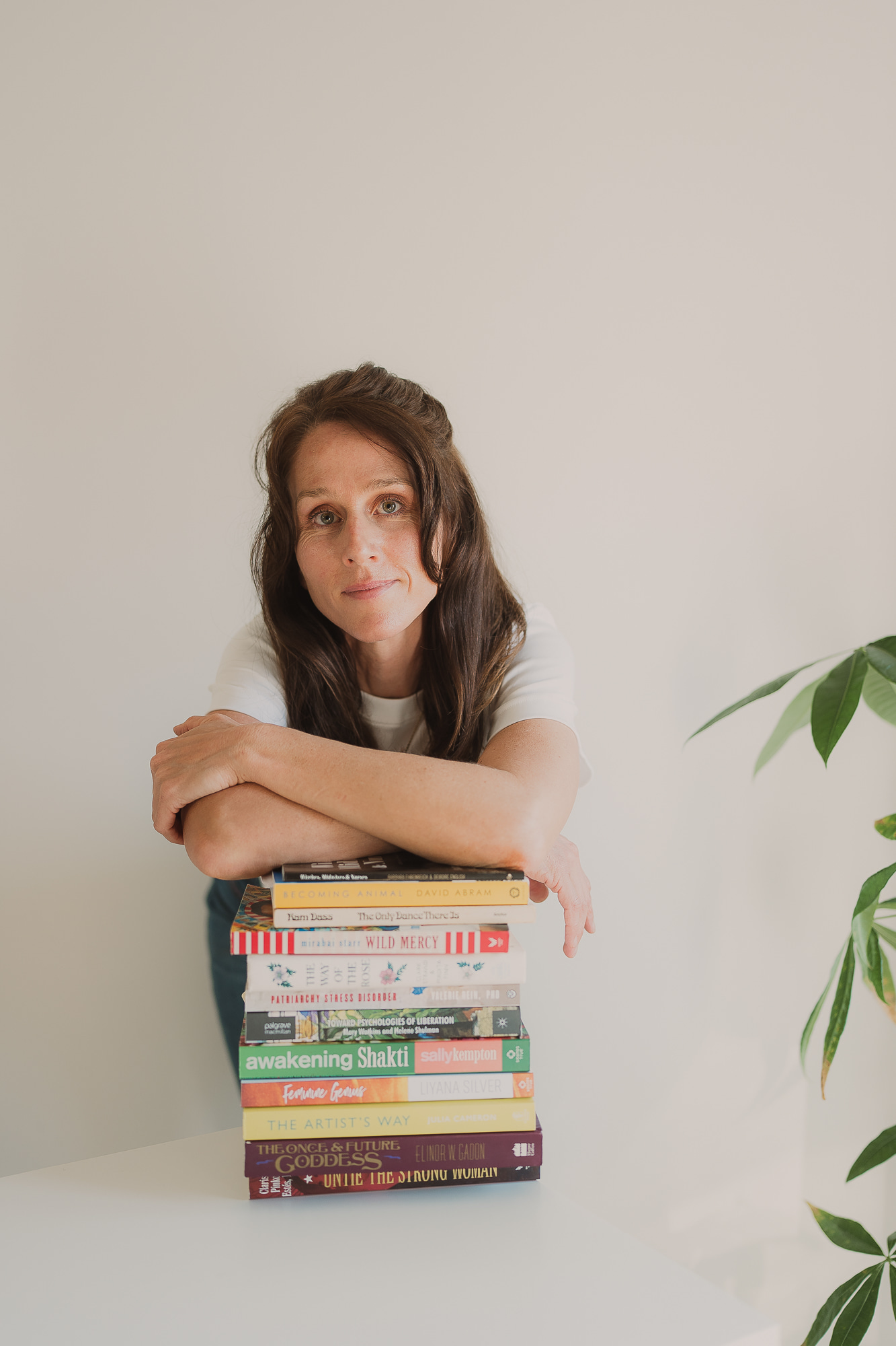 Amelia gazes at the camera, elbows resting on a stack of books on feminist psychology and feminine spirituality with a neutral background and plants in foreground