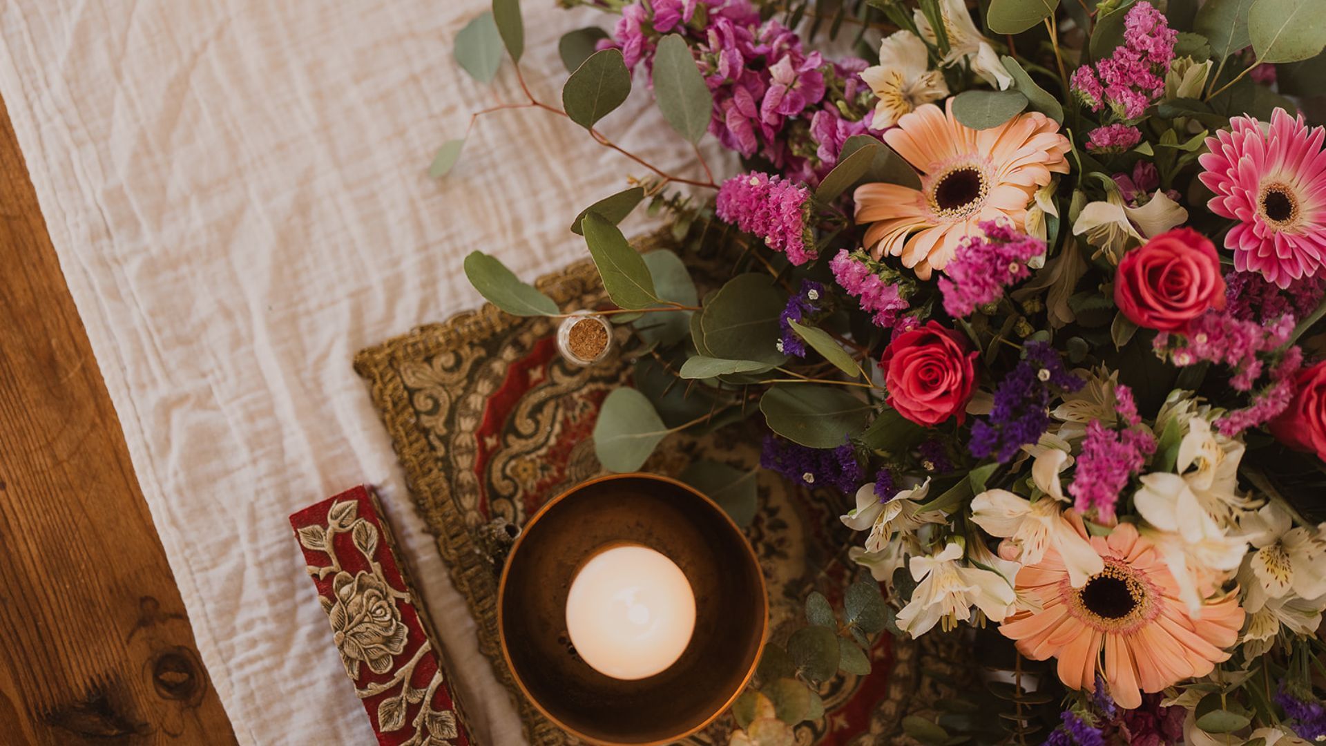 Image: Overhead shot of abundant flowers, singing bowl with candle, embroidered textile, wooden table