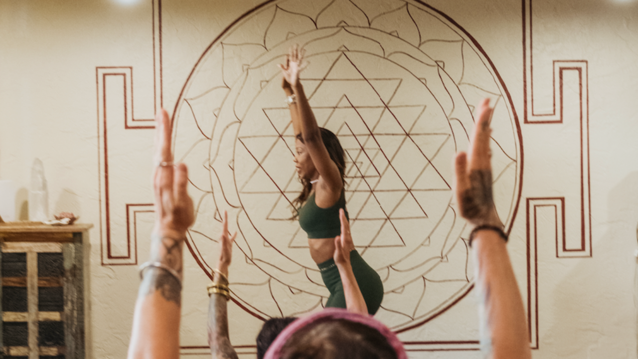 Black woman teaching yoga class in Chair Pose with hands up, standing in front of a white wall with a Sri Yantra painted in red.