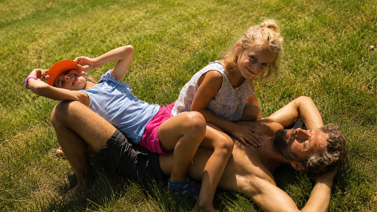 Papà sdraiato sull’erba che respira in modo naturale con due bambine che giocano sopra di lui, simbolo di corpo libero e respiro naturale
