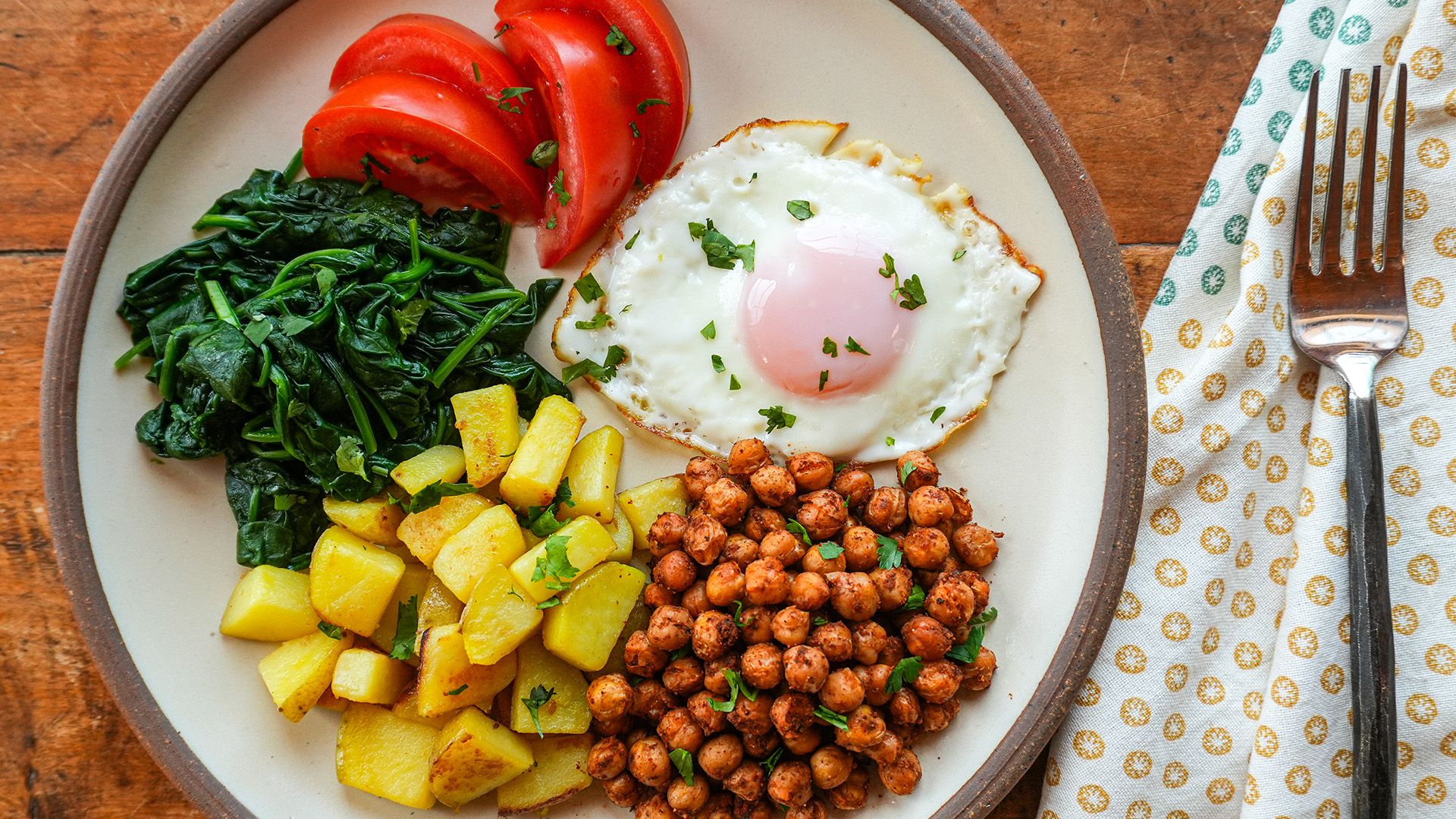 A breakfast plate featuring smoky chickpeas, diced potatoes, sautéed spinach, a sliced tomato, and a sunny-side-up egg garnished with fresh cilantro.