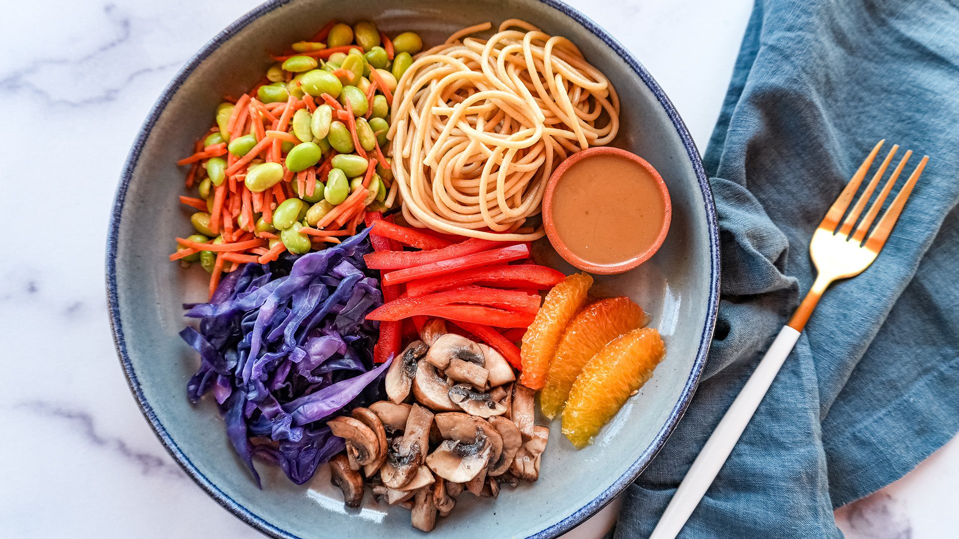 A colorful bowl of Thai peanut noodle salad topped with shredded purple cabbage, edamame, and orange segments.