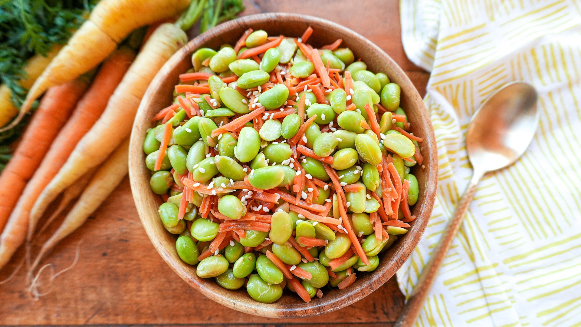 A ceramic bowl filled with a colorful edamame stir-fry featuring green soybeans, shredded carrots, and sesame seeds.