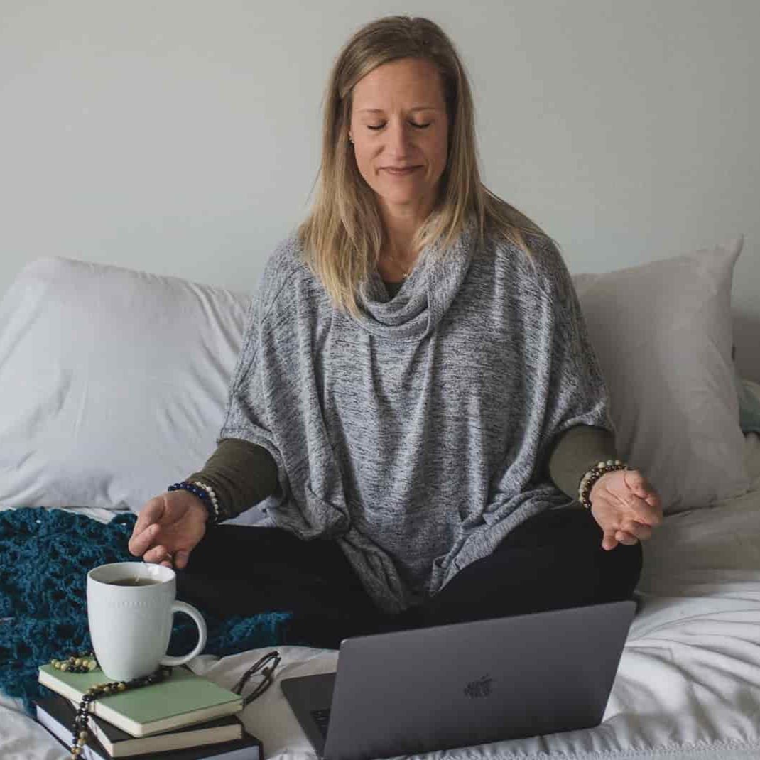 Woman sitting cross-legged on a bed in meditation, eyes closed, with a laptop in front of her and a cup of tea resting on a stack of books.