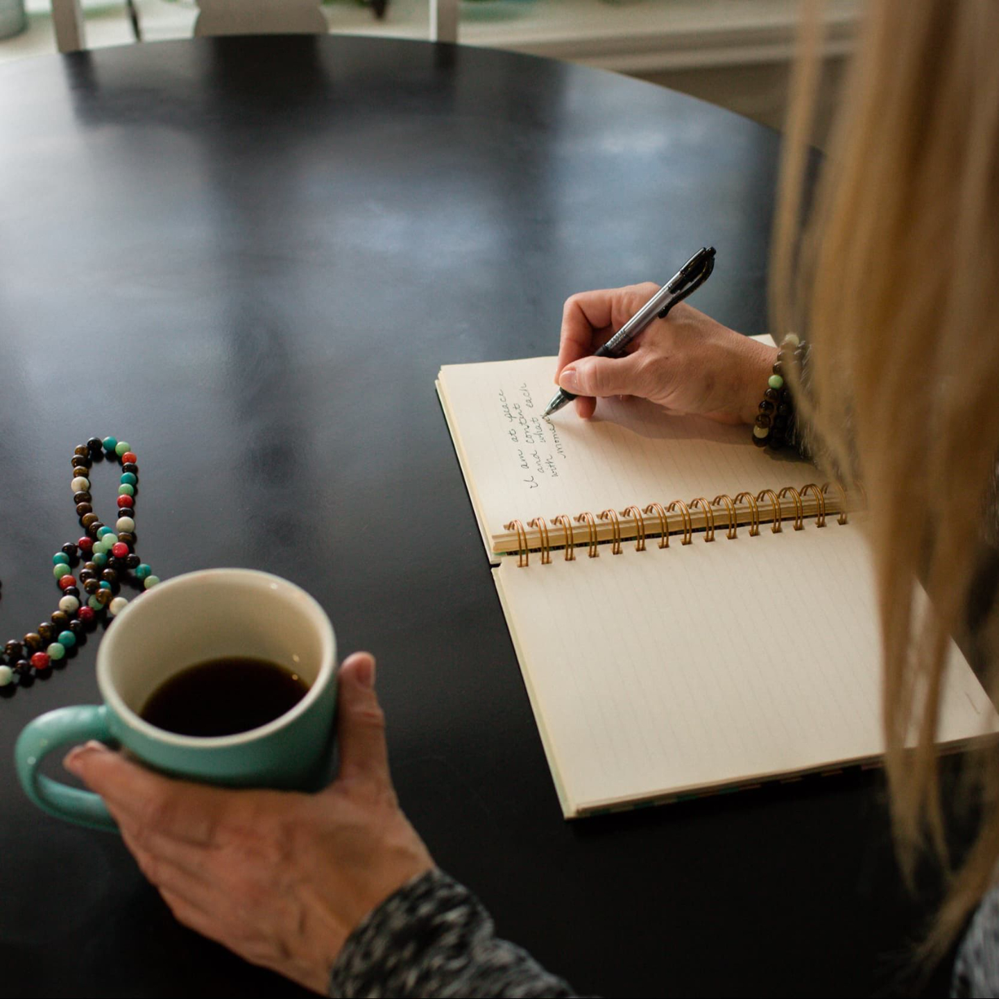 Woman journaling with coffee and mala beads on a black table near a window.