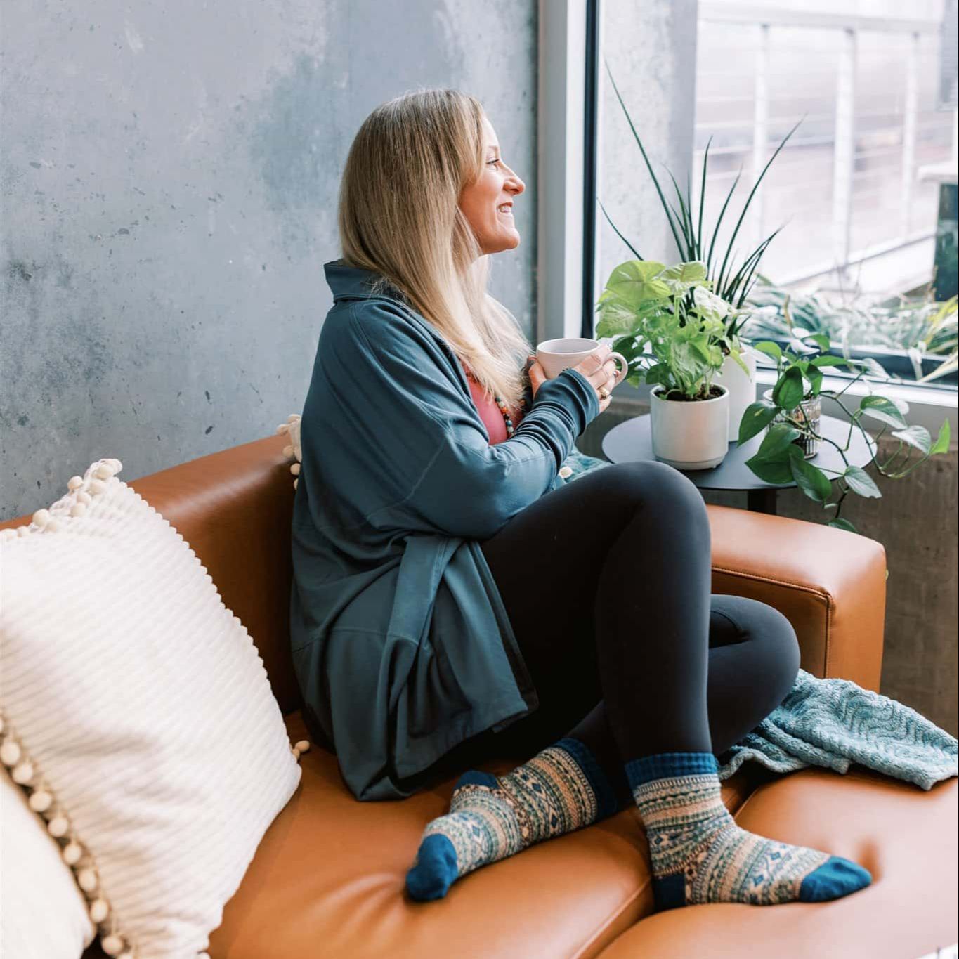 Woman sitting on a cozy leather sofa by the window, holding a mug, wearing a blue sweater and patterned socks, surrounded by houseplants.