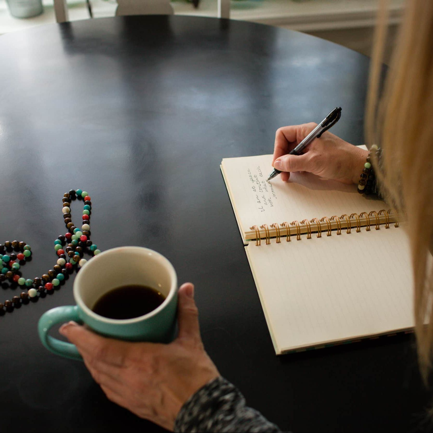 Close-up of a woman journaling at a round black table with a cup of coffee and mala beads resting beside her notebook.