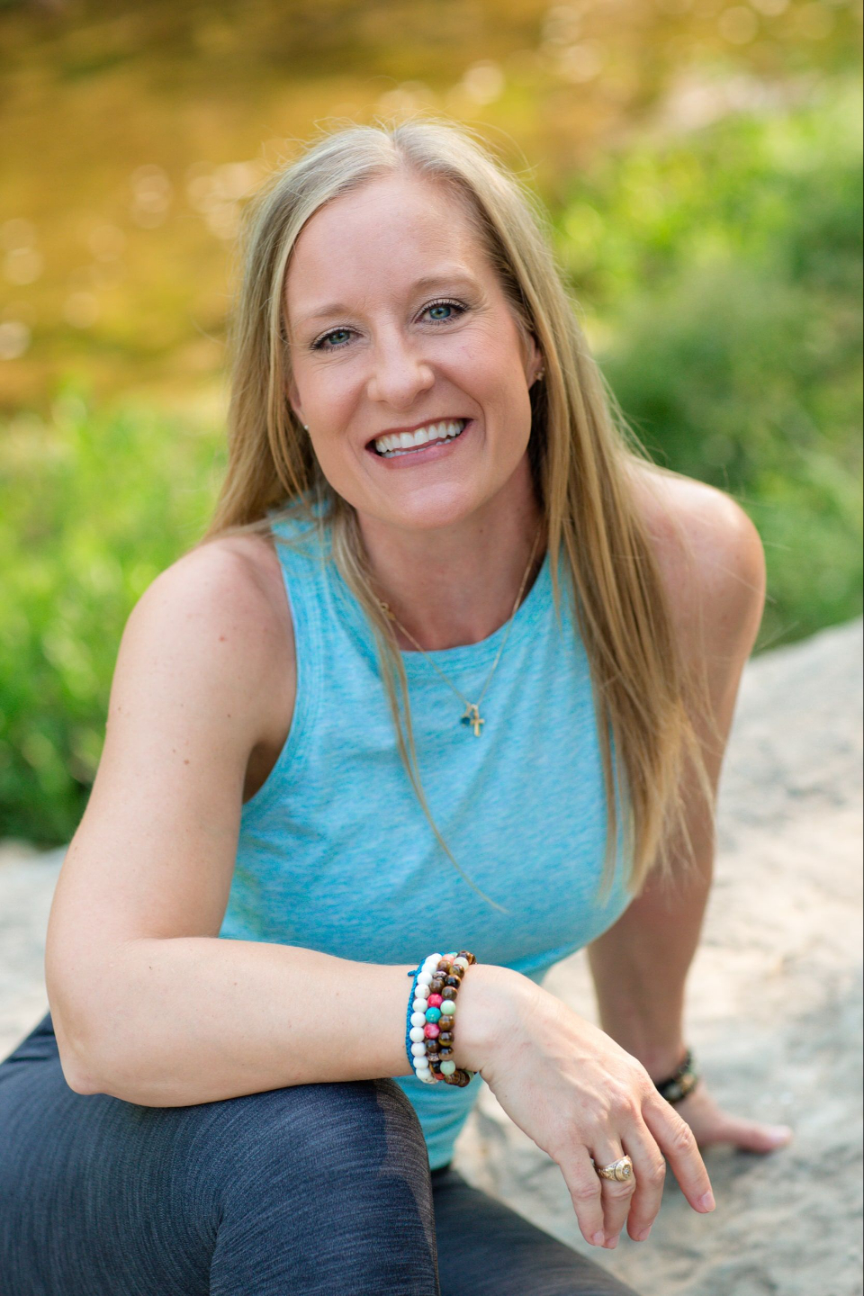 Smiling woman with long blonde hair wearing a blue top, sitting outdoors with greenery in the background.