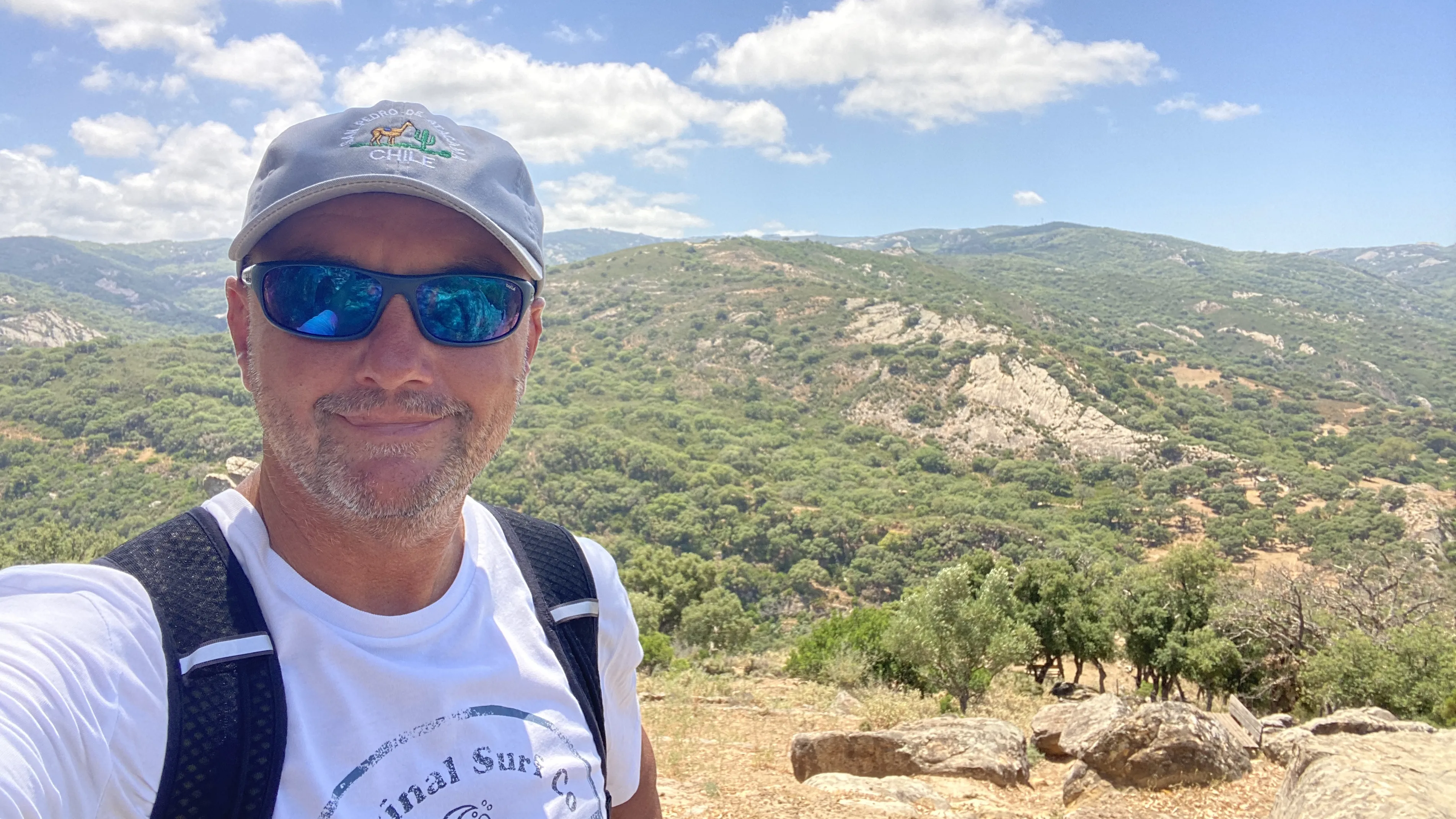 Andy on a hilltop hike in the Andalucían countryside, wearing a cap, sunglasses and a backpack, with green hills and blue sky in the background.