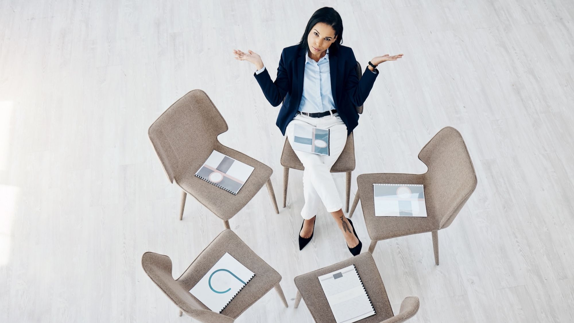 Businesswoman shrugging alone among empty chairs in a circle, viewed from above
