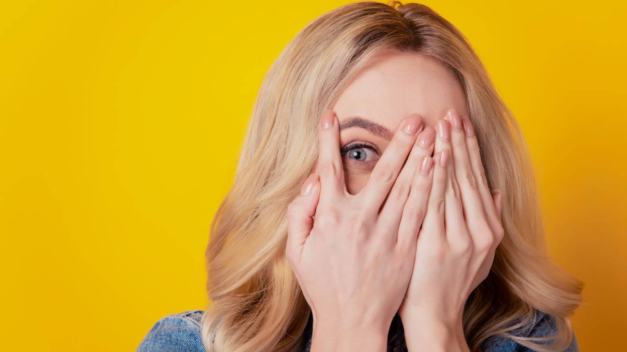 Woman peeking through her fingers against a yellow background.