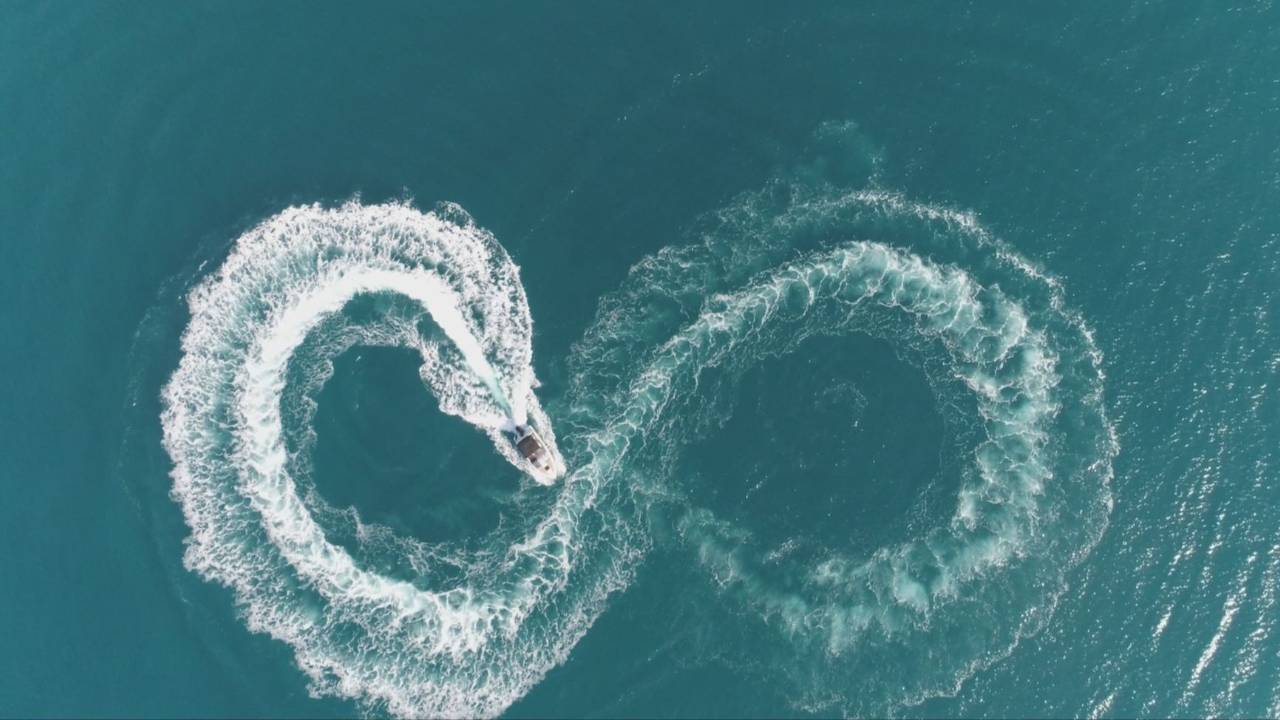 Top-down aerial shot of a boat creating a white infinity symbol wake in teal ocean waters.