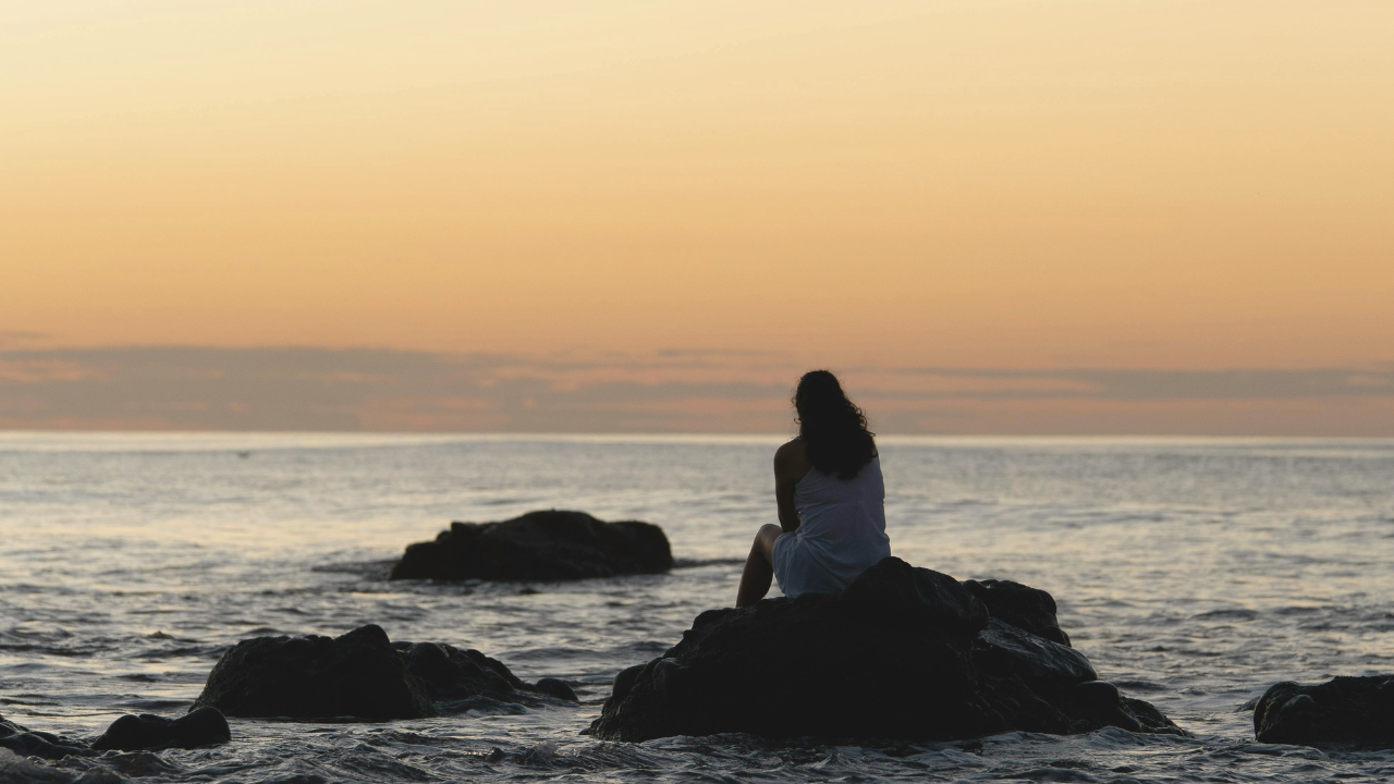 A woman sits alone on a rock in the ocean. 