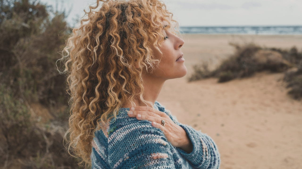 A woman stands on the beach with eyes closed and hands on her shoulders in a moment of presence. 