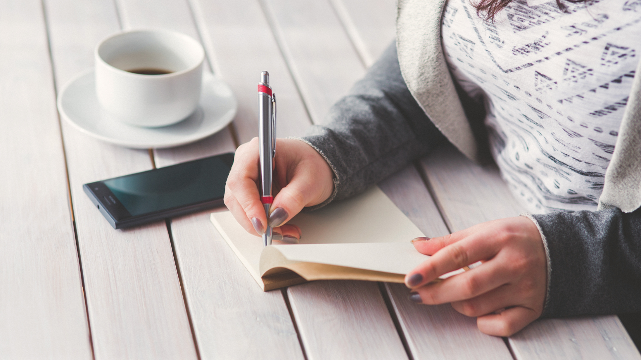 A woman sits with a journal and cup of coffe at a desk.