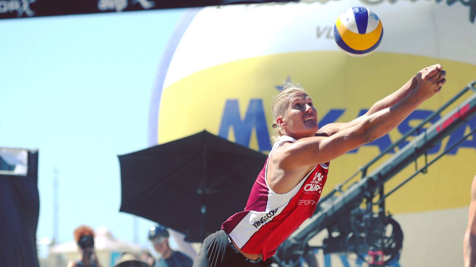 Beach volleyball player mid-air hitting a jump serve with the ball overhead at a tournament