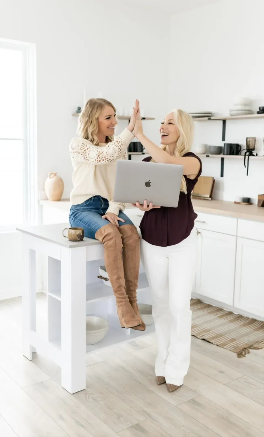 Two women high-fiving and laughing in a bright white kitchen, one seated on a table holding a MacBook laptop