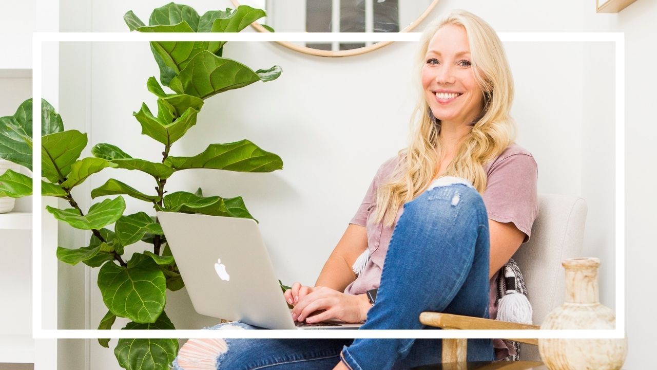 Woman smiling while working on a MacBook laptop, seated casually in a bright home office space with a fiddle leaf fig plant in the background